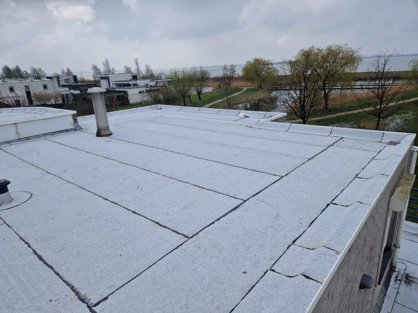 Plat dak met witte bitumen dakbedekking, ventilatiepijp en meerzicht op bomen